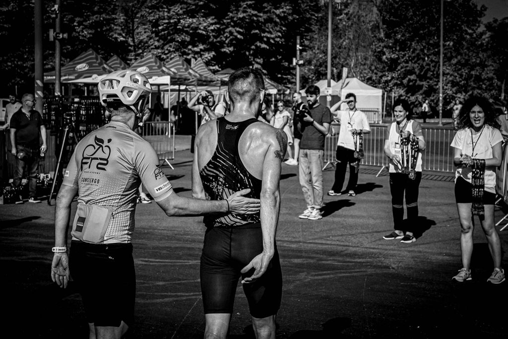 Fotografie sportiva la linia de final a Crosul Arenelor 2025: doi alergători surprinși din spate într-un gest de incurajare, voluntari care aplauda cu medaliile pregatite si fotografi documentand sosirea