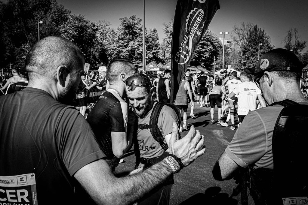 Grup de alergatori care se felicita si se incurajeaza dupa finish-ul Crosul Arenelor 2025, cu steagul oficial al evenimentului in fundal - fotografie alb-negru