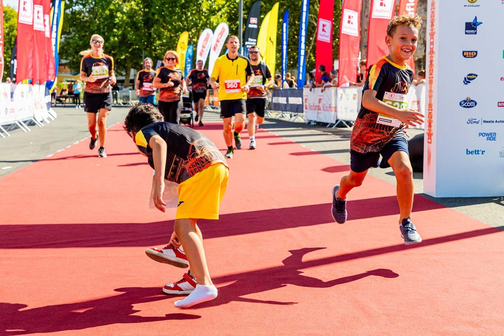 Copii pe covorul rosu la finish-ul Crosul Arenelor 2025: unul alearga zambind spre linia de sosire, iar celalalt se apleaca sa isi scoata adidasul, in timp ce alergatorii cu numere de concurs sosesc in fundal