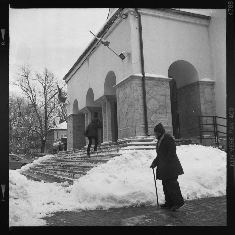 Fotografie alb-negru cu o persoană varstnică cu baston mergand pe langa o cladire cu arcade acoperita de zapada, in timp ce o alta persoana urca scarile.