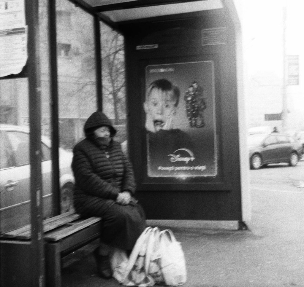 A person wearing a hooded winter coat sits on a bus stop bench with bags on the ground, while a Home Alone movie poster with the iconic surprised face is displayed on the shelter wall behind them in a bus station in Bucharest, Romania.
