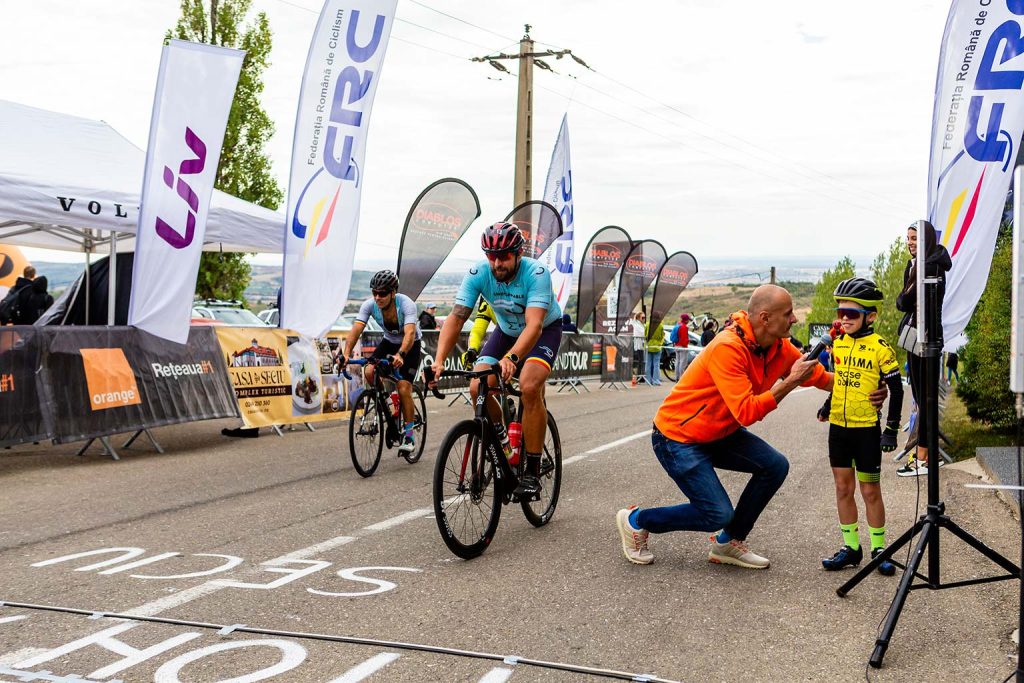 A cyclist in a blue jersey crosses the finish line while a man in an orange jacket kneels to interview a young child in a yellow cycling jersey on the sidelines at the finish line of Seciu Mon Amour road bike race in Romania.