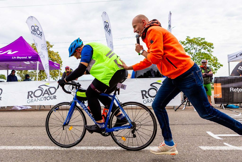 Alex Ciocan hoodie holding a microphone pushes a cyclist wearing a neon yellow vest and blue helmet on a blue bicycle for one more lap.