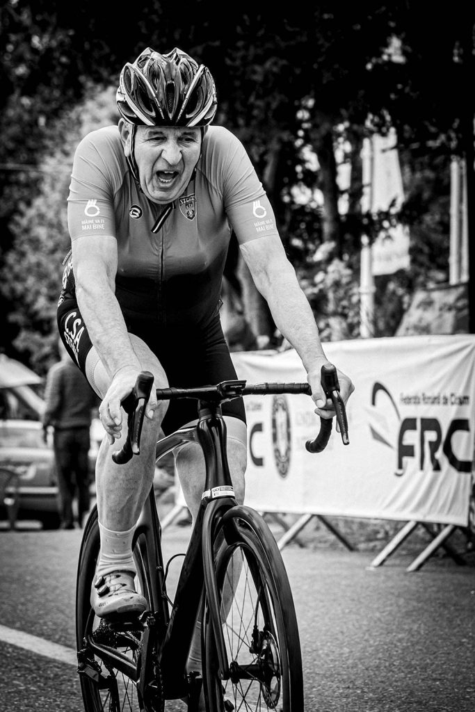 A black and white photo of an older male cyclist leaning forward on a road bike with an intense expression, wearing a helmet and racing jersey.