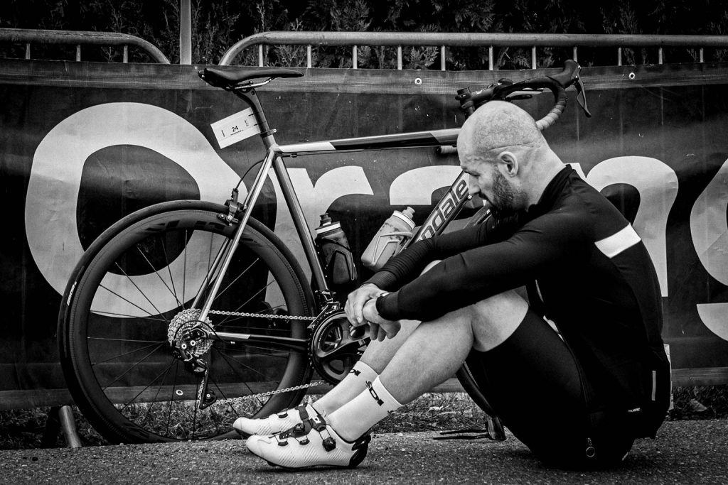 A bald cyclist in black cycling gear and white cycling shoes sits on the ground beside his road bike with race number 24, resting against a barrier in this black and white photograph.
