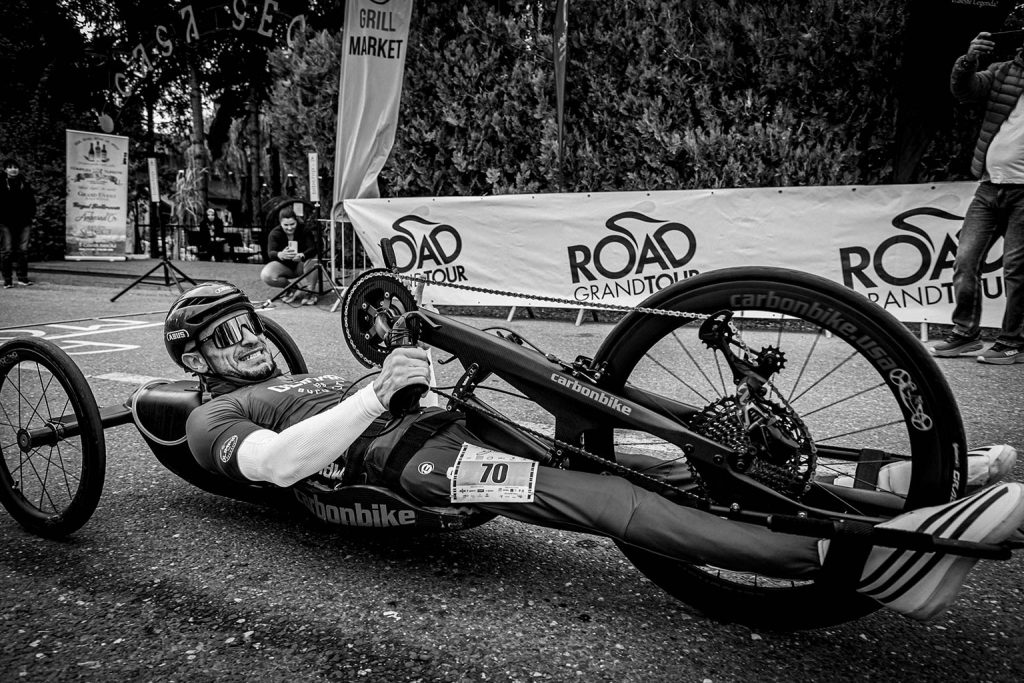 A para-cyclist in a recumbent racing bike at a ROAD GRAND TOUR event, wearing helmet and sunglasses with race number 70.