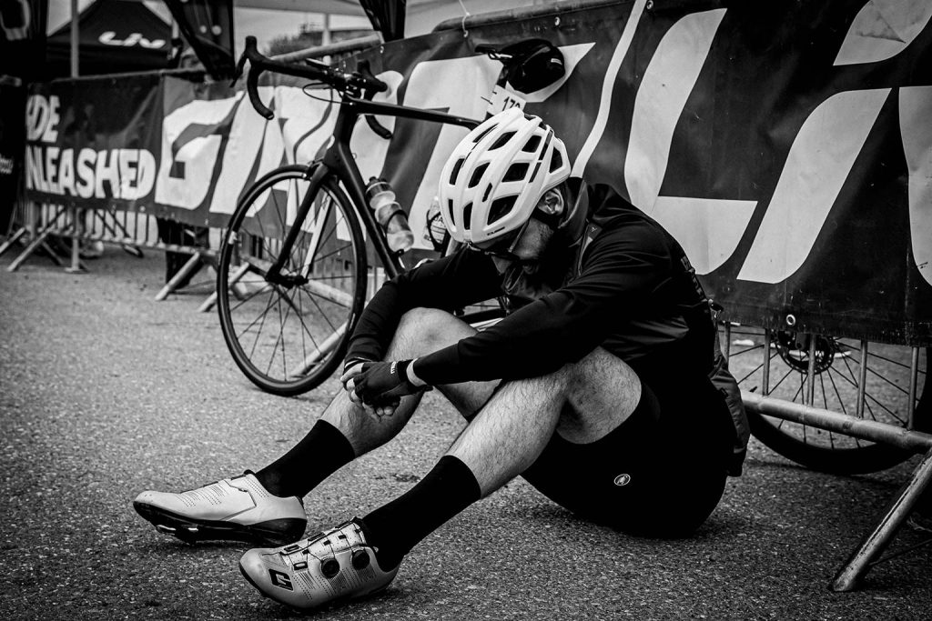 A black and white photograph of a cyclist wearing a white helmet and cycling gear, sitting exhausted on the pavement next to their road bike after finishing Seciu Mon Amour bike race.