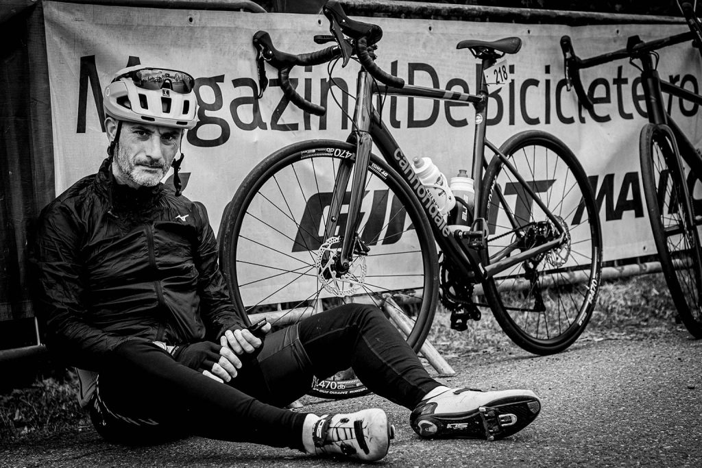 Cyclist wearing helmet and black cycling gear sitting on ground resting beside racing bicycles at cycling event, black and white photograph