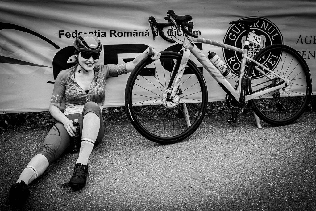 A black and white photo of a smiling female cyclist in full gear sitting on the pavement next to her white road bike, holding the front wheel, with a Romanian Cycling Federation banner in the background.