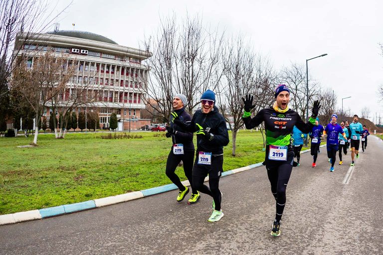 Alergatori in timpul Semimaratonului Gerar 2026 de-a lungul unui drum pavat marginit de copaci, în fundal, cladirea Rectorului UNSTPB (POLITEHNICA Bucuresti).