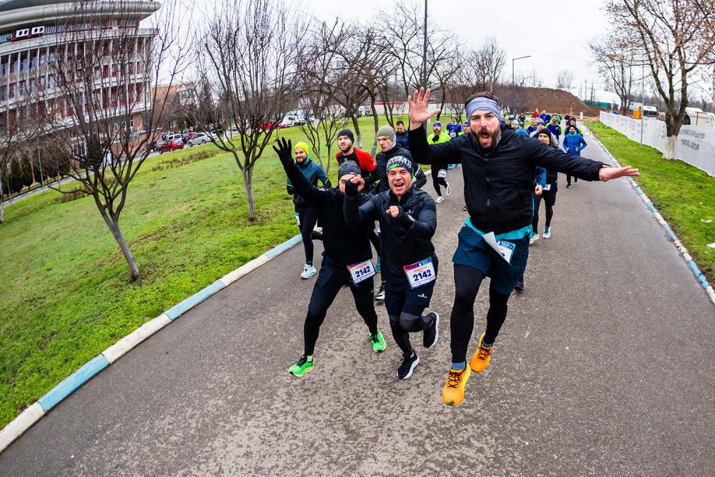 Runners during Semimaraton Gerar 2026 along a paved road lined with trees, in the background, the UNSTPB (POLITEHNICA Bucharest) Rector's Office building.