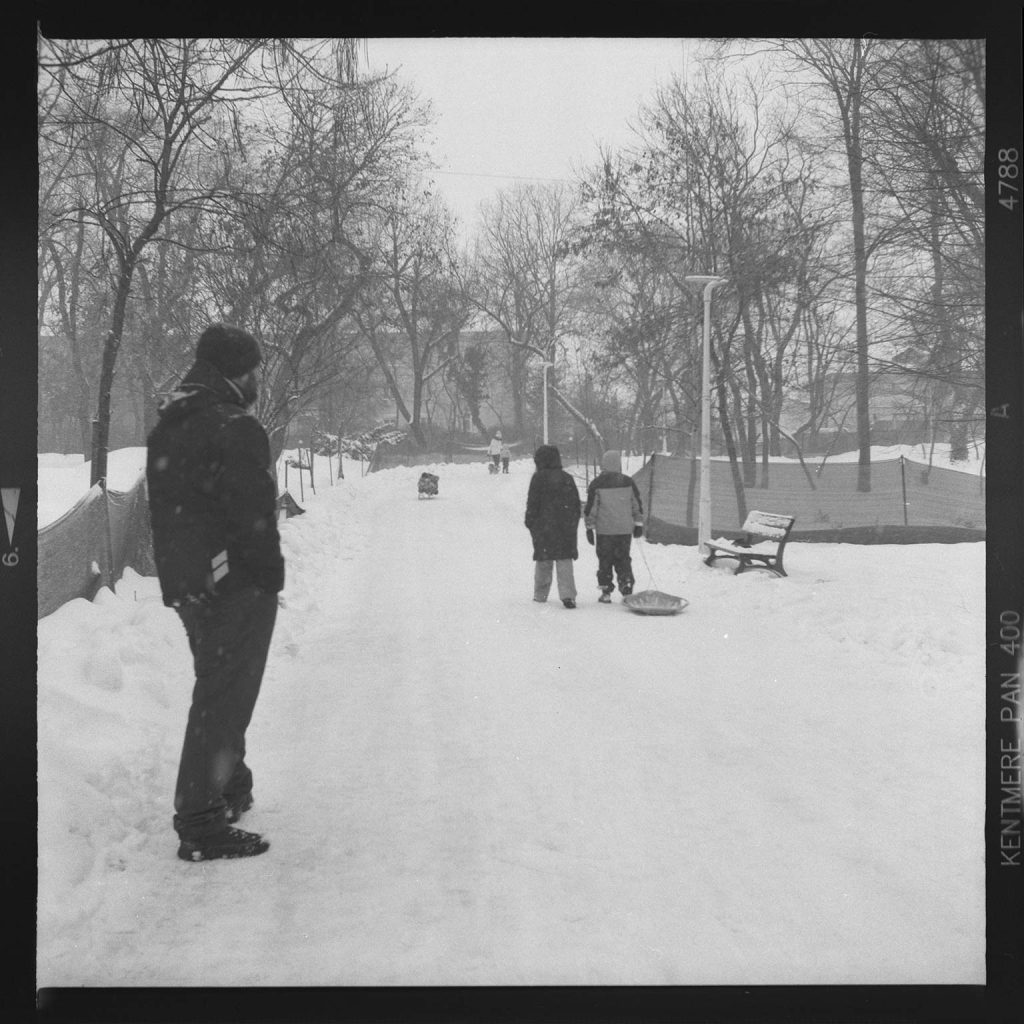 A man in a coat watches two children walk away on a snow-covered path. One child pulls a sled as they head towards bare trees under an overcast sky.
