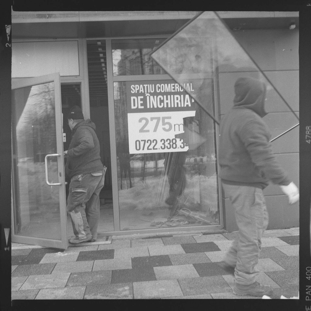 Black and white street photography showing two construction workers in hoodies and work clothes at a commercial building entrance in Bucharest, Romanian