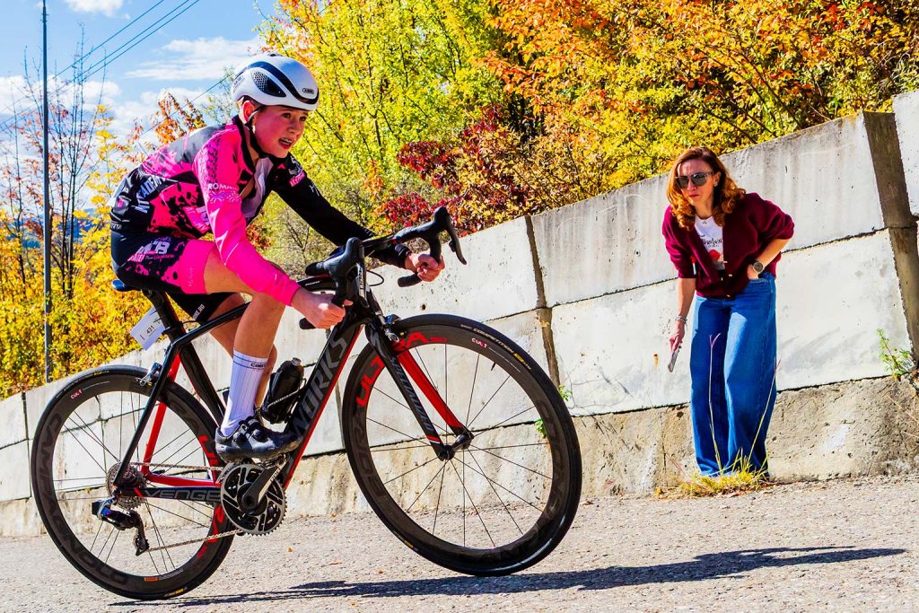 Tanar ciclist in echipament roz si negru pe o bicicleta S-Works urcand Dealul Sultanul in timpul cursei The Wall 2025, inconjurat de peisaj de toamna si urmarit de o spectatoare care il incurajeaza