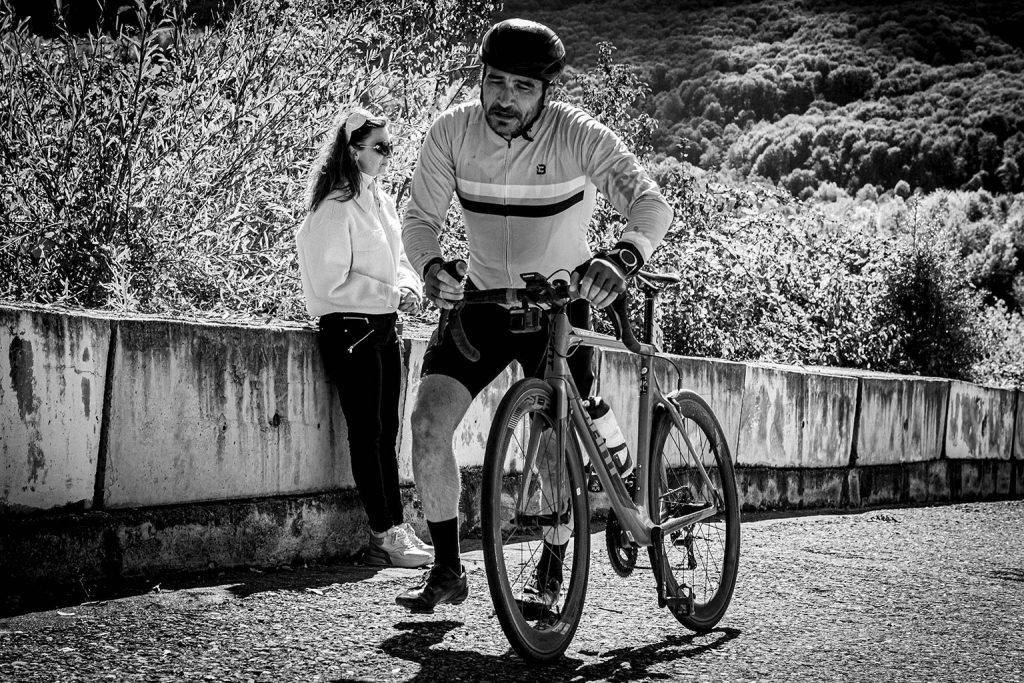 Ciclist impingandu-si bicicleta in timpul urcarii pe Dealul Sultanul in timpul cursei The Wall 2025, in timp ce o spectatoare priveste de pe margine. Fotografie alb-negru.