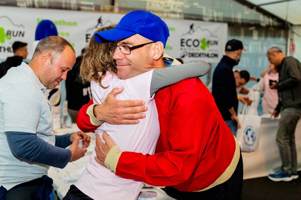 Barbat cu sapca albastra si geaca rosie imbratisand o voluntara la standul de inscrieri Eco Run 2026 organizat de Decathlon.