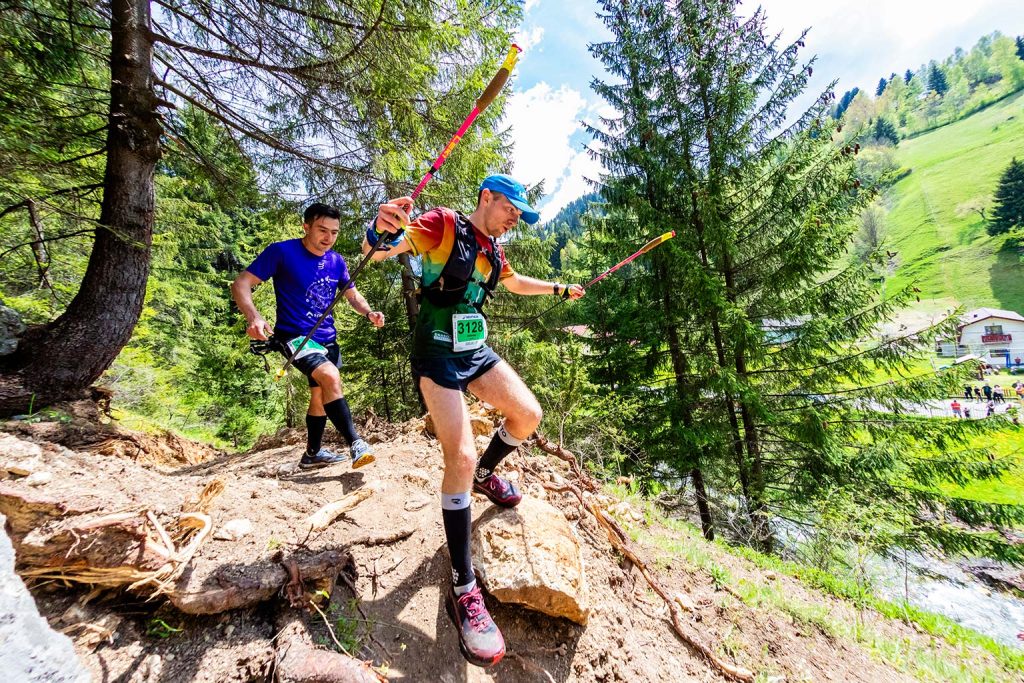 Concurent cu numarul 3128 si bete de trekking coborand rapid pe un traseu abrupt in padure la EcoRun Moieciu 2026, fotografiat cu obiectiv fish eye.