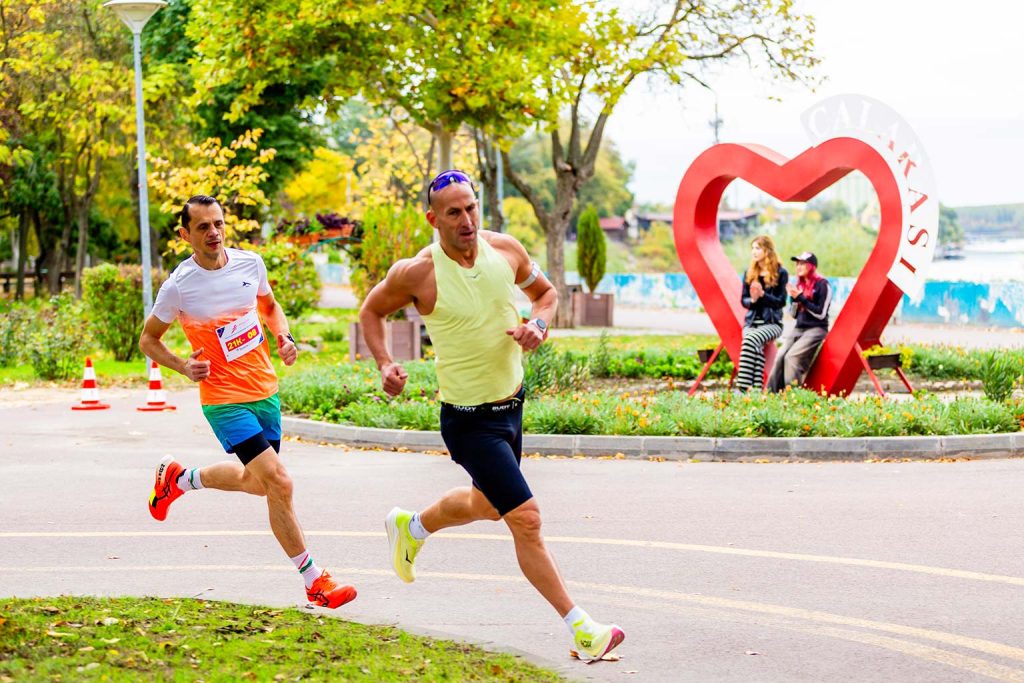 Doi alergatori barbati in fruntea cursei de semimaraton, trecand pe langa sculptura in forma de inima rosie cu inscriptia Calarasi.