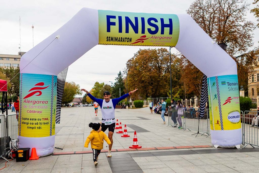 Concurent trecand sub arcul de finish cu bratele deschise, alaturi de un copil in haina galbena, la Semimaratonul Alergarea de Sambata Calarasi.