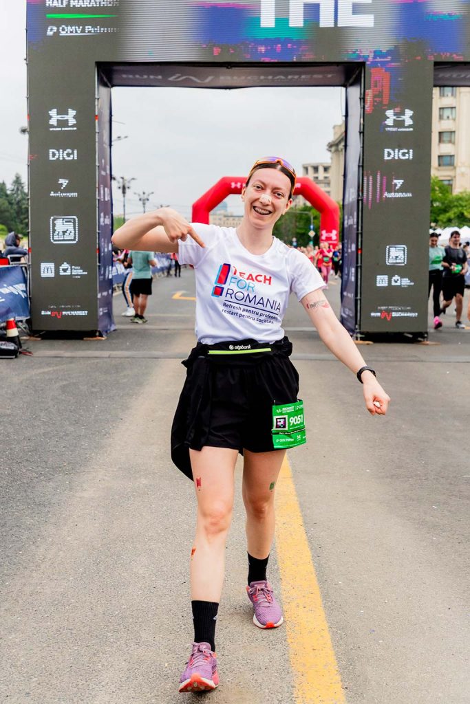 Alergatoare zambitoare cu tricou Teach For Romania aratand spre logo in fata arcului de finish la OMV Petrom Bucharest Half Marathon 2026.