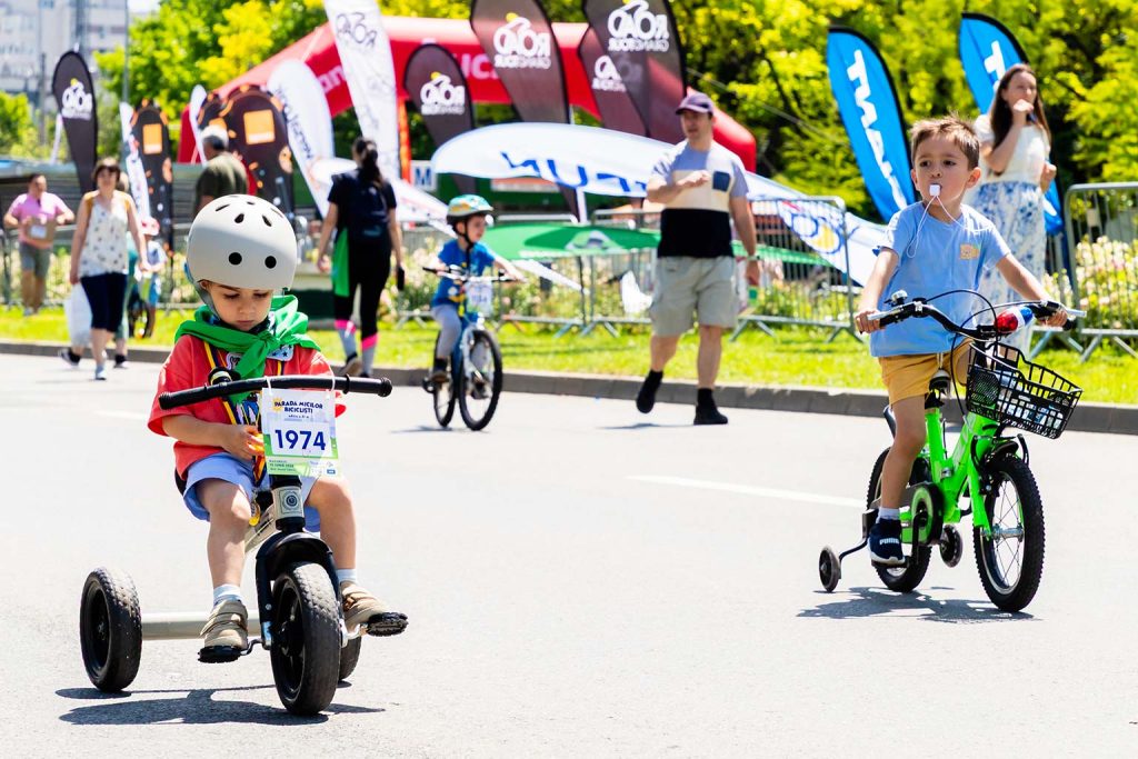 Doi copii pe biciclete cu rotite ajutatoare la Parada Micilor Ciclisti, participant cu numarul 1974 si copil pe bicicleta verde