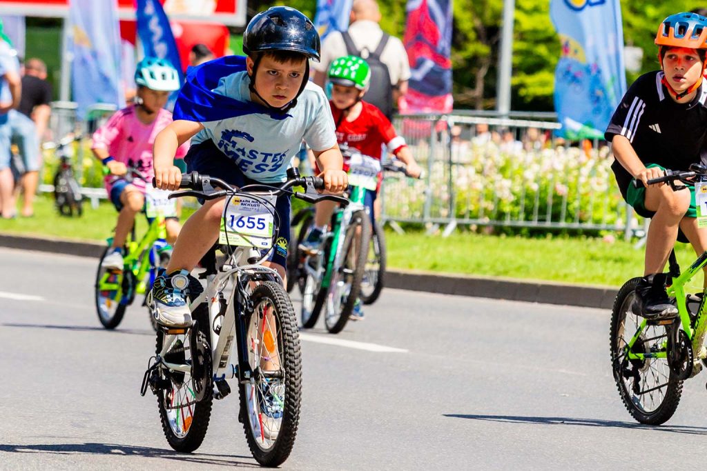 Copil cu pelerina albastra si numarul 1655 pedaland concentrat in Parada Micilor Ciclisti, urmat de alti participanti
