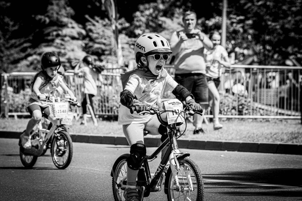 Copil cu ochelari de soare si casca, exprima entuziasm pe bicicleta in timpul Paradei Micilor Ciclisti, imagine alb-negru
