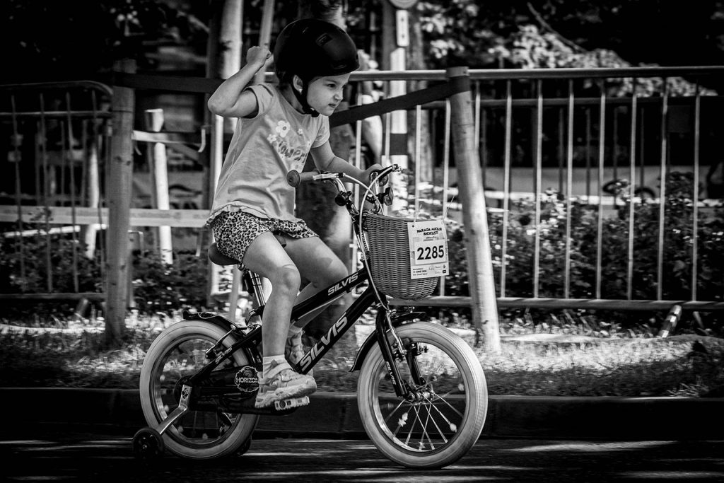 Copil cu numarul 2285 pe bicicleta cu cosulet si rotite ajutatoare, ridicand pumnul in aer la Parada Micilor Ciclisti, imagine alb-negru