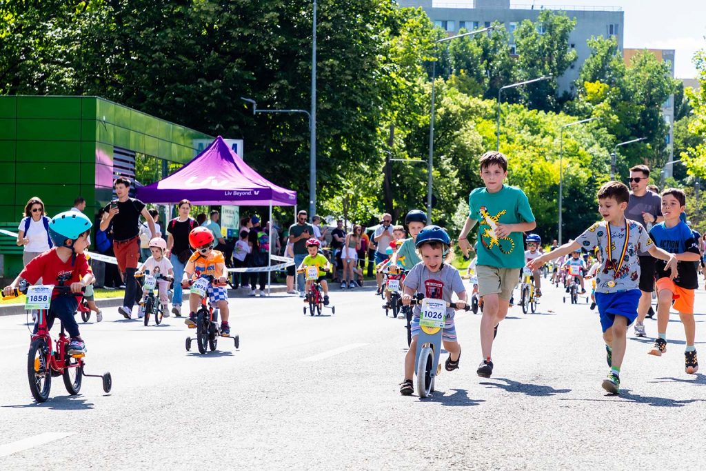 Grup de copii participand la Parada Micilor Ciclisti, unii pe biciclete cu numar de concurs, altii alergand, intr-o zi insorita