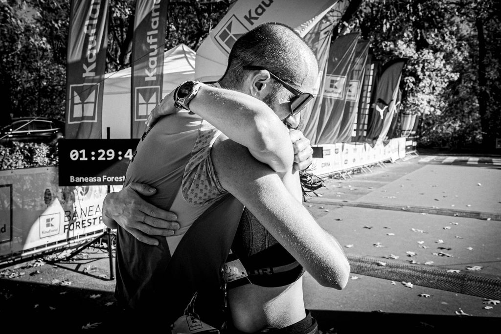 Doi alergatori imbratisandu-se emotional la linia de finish a Baneasa Forest Run 2025, cu cronometrul in fundal, fotografie alb-negru
