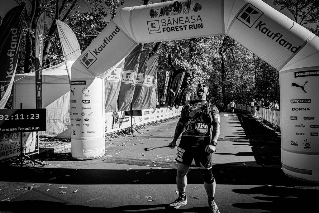Alergator barbat urland de triumf sub arcada Baneasa Forest Run 2025, cronometru 02:11:23, fotografie alb-negru dramatica
