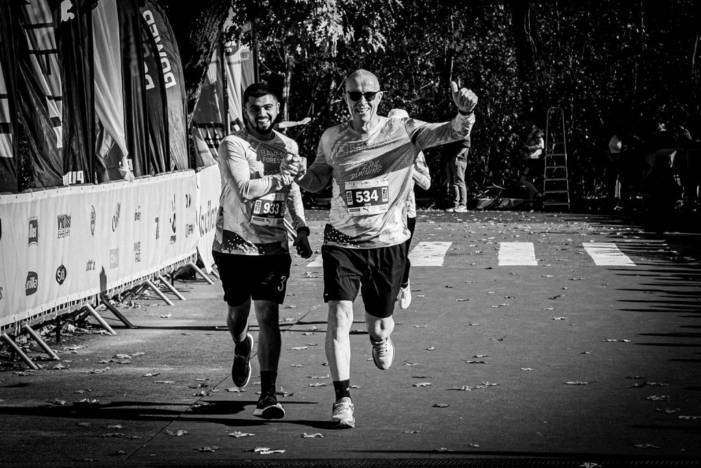Doi alergatori zambind si tinandu-se de mana la finishul Baneasa Forest Run 2025, numere 933 si 534, fotografie alb-negru