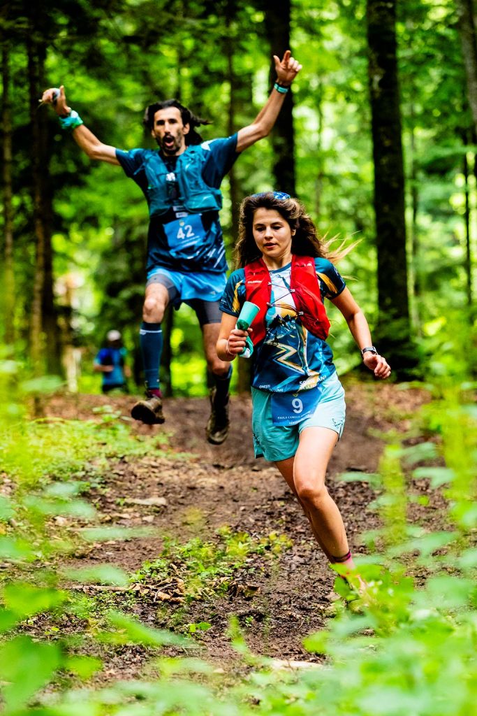Legendele Nemirei - fotografie sportiva - Doi alergatori pe traseu montan in padure, barbatul cu numarul 42 sarind cu bratele ridicate, femeia cu numarul 9 alergand in fata, echipament sportiv albastru, natura salbatica.