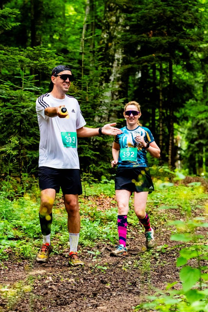 Legendele Nemirei - fotografie sportiva - Doi alergatori participand la cursa Legendele Nemirei in padure, barbatul cu numarul 393 in tricou alb Adidas si ochelari de soare, femeia cu numarul 392 in echipament albastru-verde, sosete colorate roz-albastru, traseu montan printre copaci.