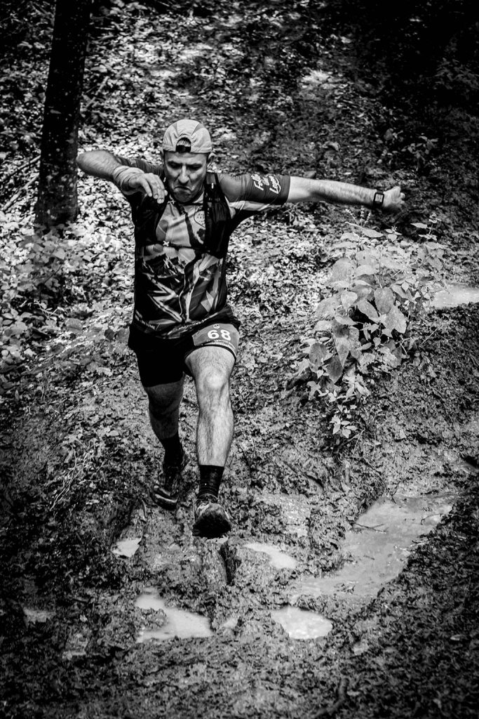 Legendele Nemirei - fotografie sportiva - Alergator barbat cu numarul 68, echipat cu sapca si tricou montan, navigand prin teren noroios in padure, bratele intinse pentru echilibru, fotografie alb-negru