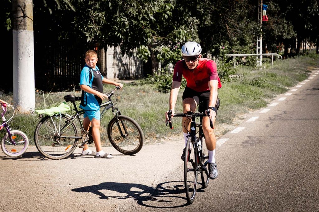 Ciclist in echipament rosu pedalând pe sosea, privit de un copil care sta cu bicicleta langa drum, in cadrul Turului Ciclist CELCO. Fotografie sportiva ciclism rutier surprinsa de Popa Bogdan.