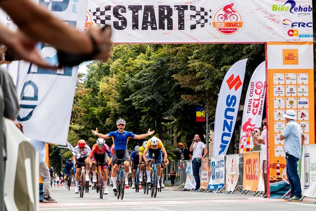 Fotografie sportiva la concurs de bicicleta sosea surprinzand castigatorul probei criterium care traverseaza linia de sosire cu bratele deschise.