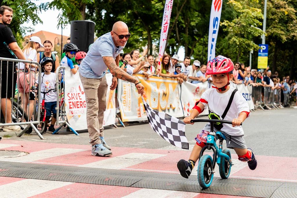 Fotografie sportiva la concurs de copii surprinzand cel mai tanar participant pe o bicicleta de echilibru trecand linia de sosire in timp ce un adult flutura steagul in final.