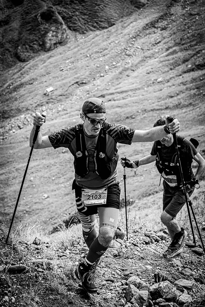 Alergator montan cu numarul 2103 urcand abrupt cu bete de trekking in Valea Ciubotea la Omu Marathon 2025, urmat indeaproape de un alt concurent - fotografie sportiva alb-negru.