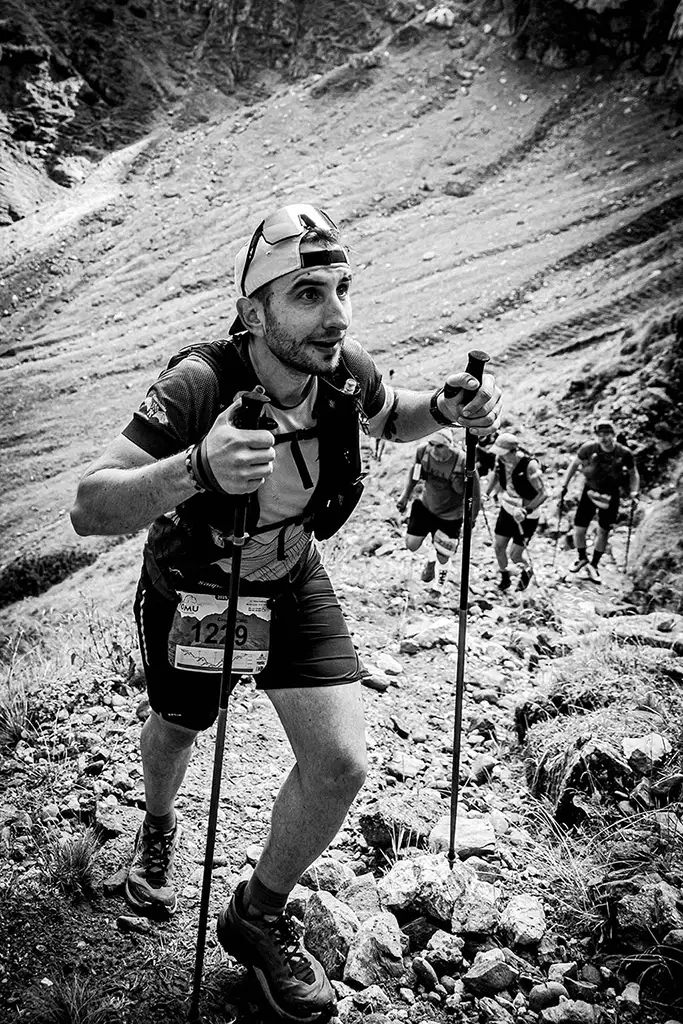 Alergator montan cu numarul 1229 urcand cu bete de trekking pe poteca pietroasa din Valea Ciubotea la Omu Marathon 2025, urmat de alti concurenti - fotografie sportiva alb-negru.