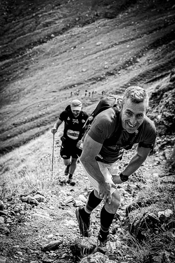 Alergator montan zambind catre camera in timp ce urca aplecat pe poteca abrupta din Valea Ciubotea la Omu Marathon 2025, urmat de alti concurenti - fotografie sportiva alb-negru.